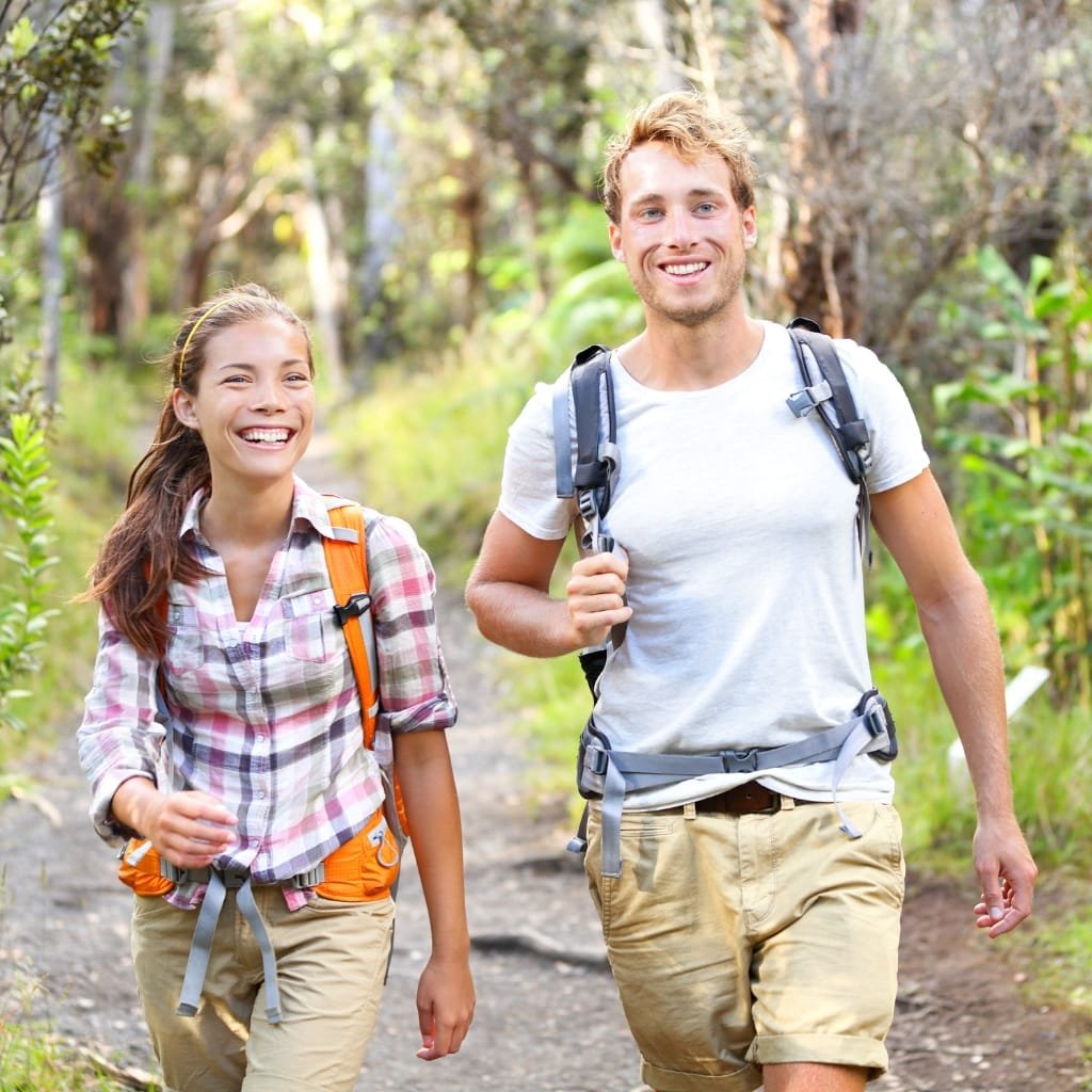 How to Style an Anorak Rain Jacket for Every Occasion 6 Two young adults, a woman and a man, smiling and enjoying a hike through a lush, green forest on a sunny day. They are carrying backpacks and wearing casual outdoor clothing.