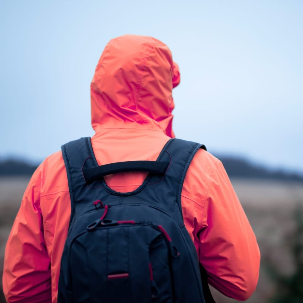 A person wearing a bright orange rain jacket with the hood up, carrying a navy blue backpack, suitable for travel in rainy weather.
