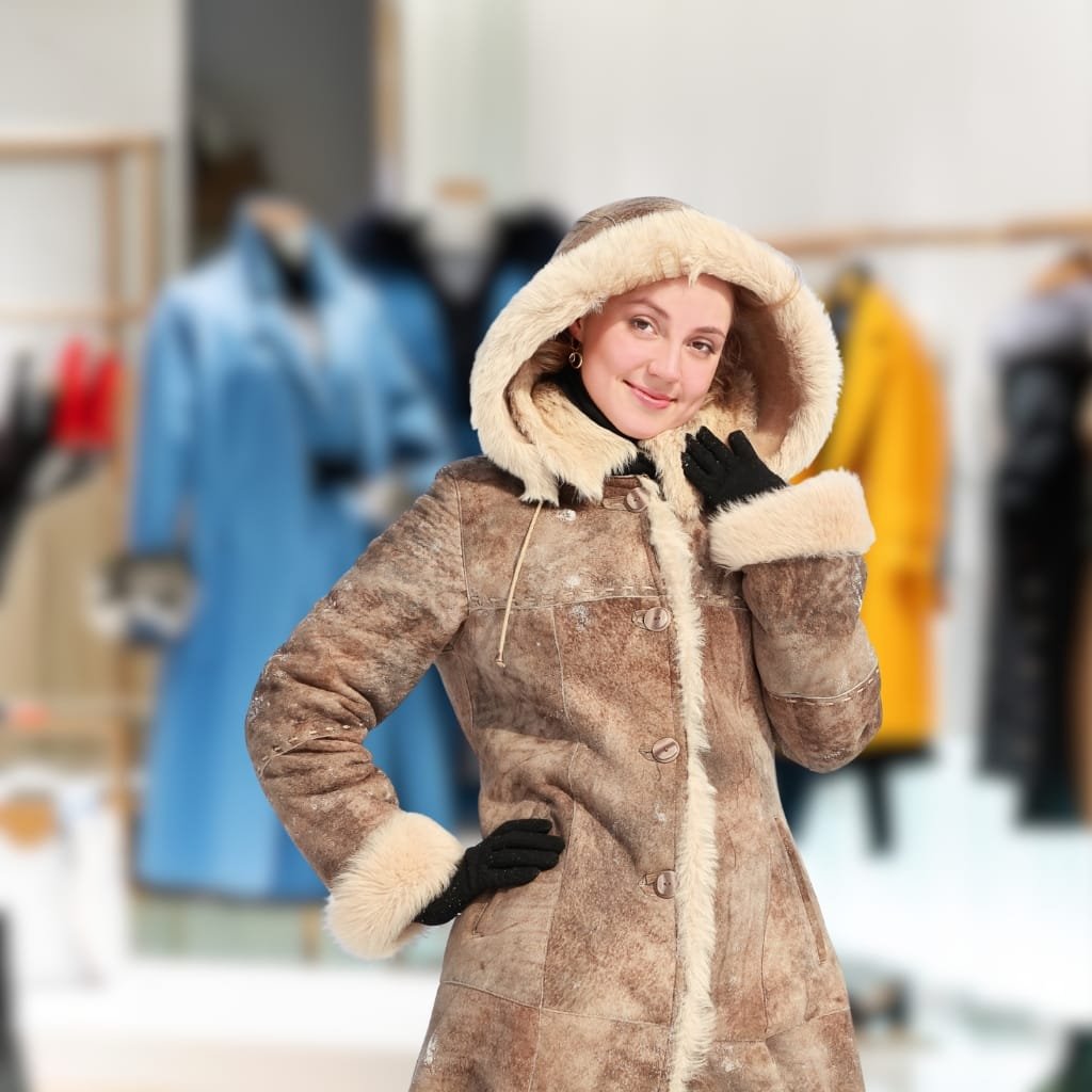 A woman in a cozy beige overcoat with a fur-lined hood, standing inside a clothing store and smiling.