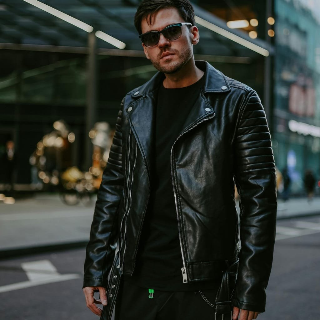 A person wearing an oversized black bomber jacket with quilted shoulder details, paired with sunglasses, standing outdoors in an urban setting.