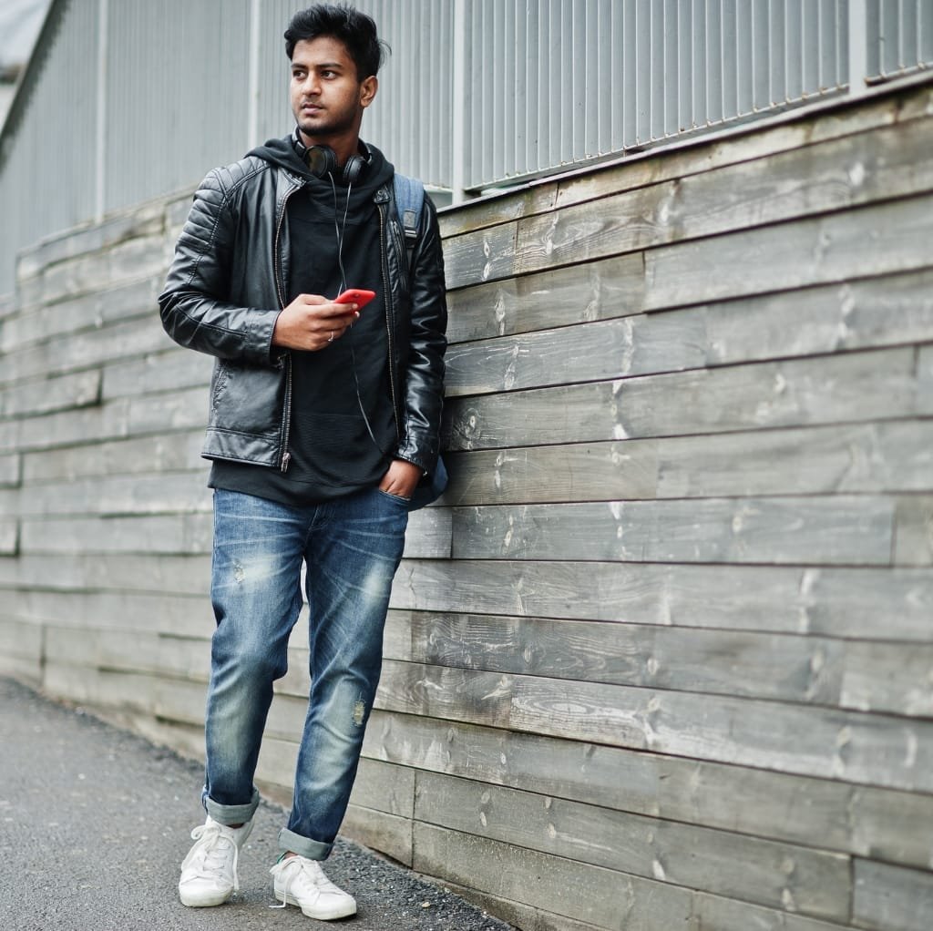 A young man standing outdoors against a wooden fence, wearing a black reversible bomber jacket paired with distressed blue jeans and white sneakers. He holds a red phone and has headphones around his neck.