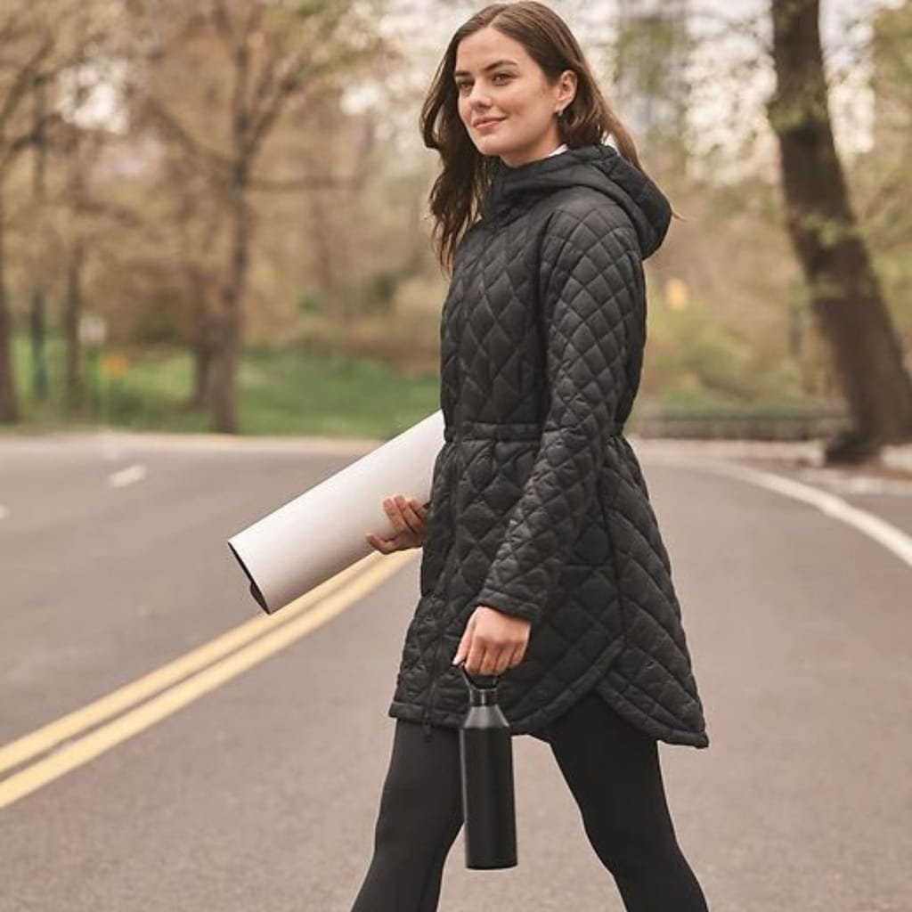 A woman walking outdoors on a paved road, wearing a black quilted parka with a hood, holding a sports water bottle in one hand and a rolled-up yoga mat in the other, with trees and autumn foliage in the background.