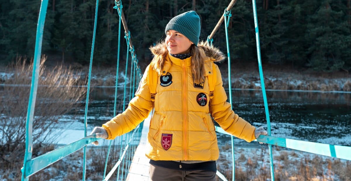 Parkas Coat Trends: Cozy Outerwear for Every Season A woman in a yellow parka coat standing on a suspension bridge over a river, dressed warmly with a blue beanie and gloves, in a winter landscape with snow and trees.