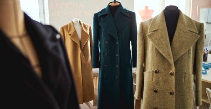 Four stylish pea coats displayed on mannequins in a well-lit room, featuring different colors and designs, with a blurred background that includes a window and some office supplies.