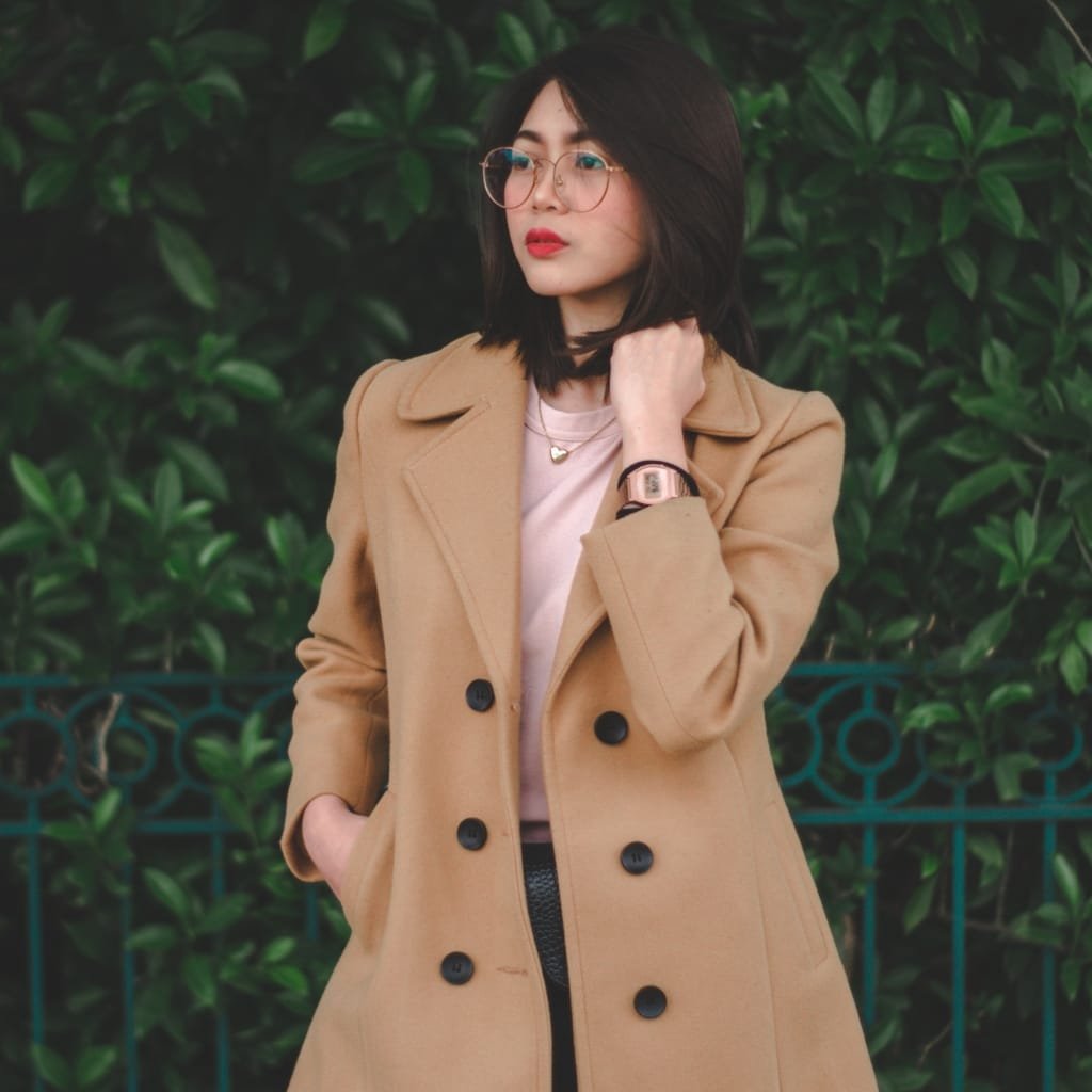 A young woman standing outdoors in front of greenery, wearing a light brown peacoat with her hand near her neck, accessorized with glasses, a watch, and delicate jewelry.

