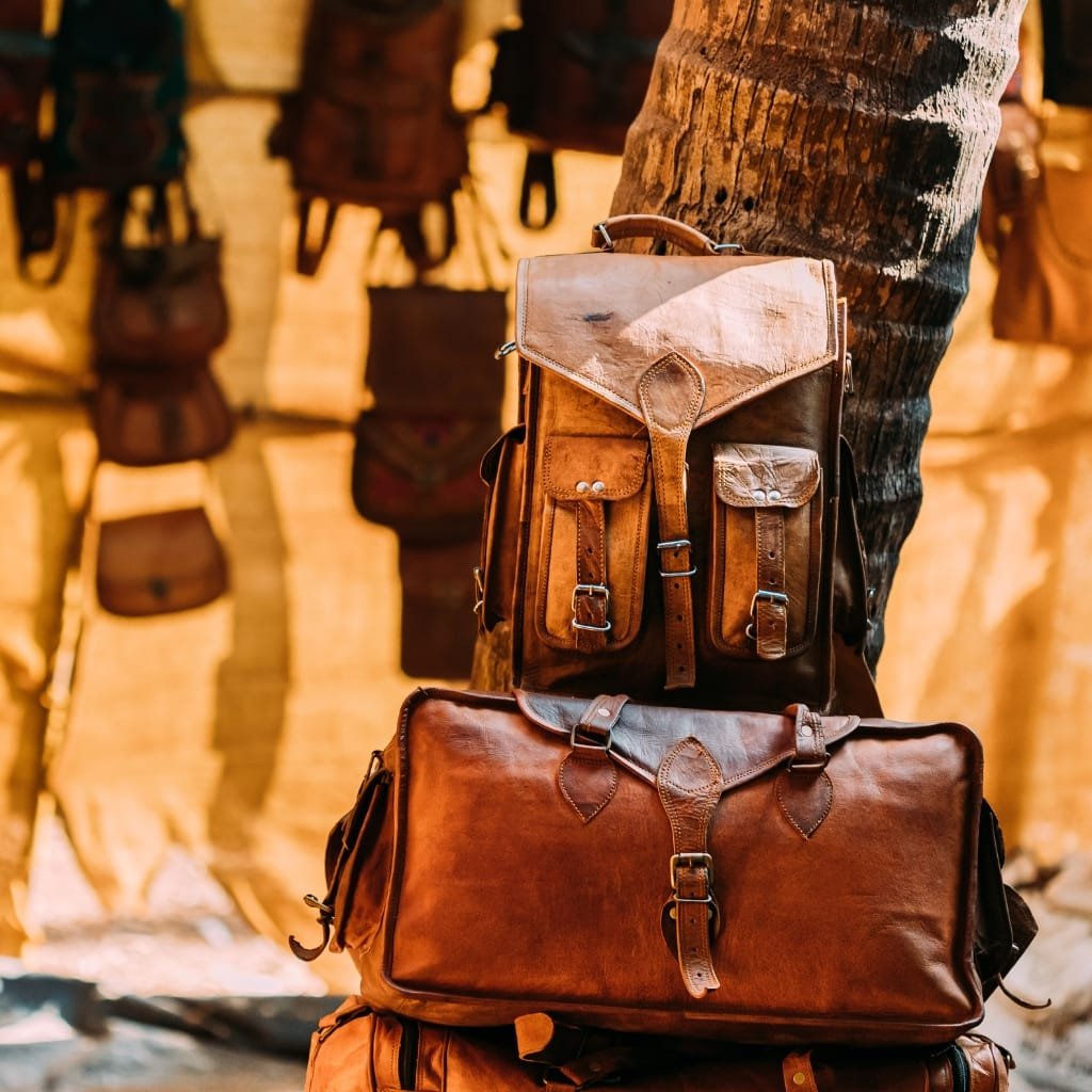 Three leather bags made of goat leather displayed in a rustic setting, with a background of hanging bags and a wooden wall.