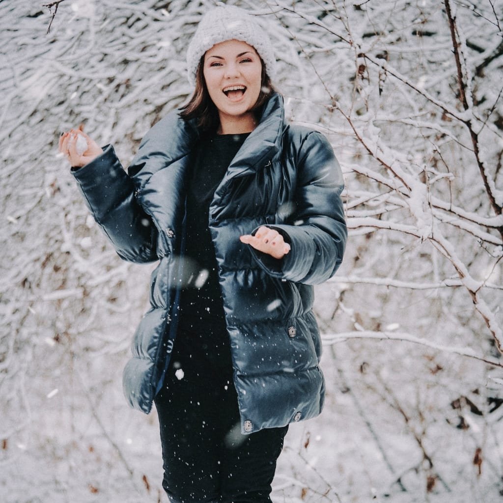 A smiling woman wearing a shiny black puffer coat and a white knitted beanie, standing in a snowy forest, enjoying winter.