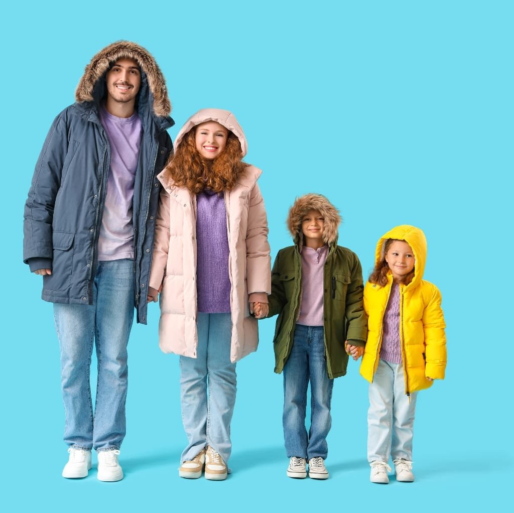 A family of four dressed in colorful winter jackets, standing against a light blue background. The father and older girl wear blue jackets, the mother and younger girl wear yellow jackets, all with hoods.