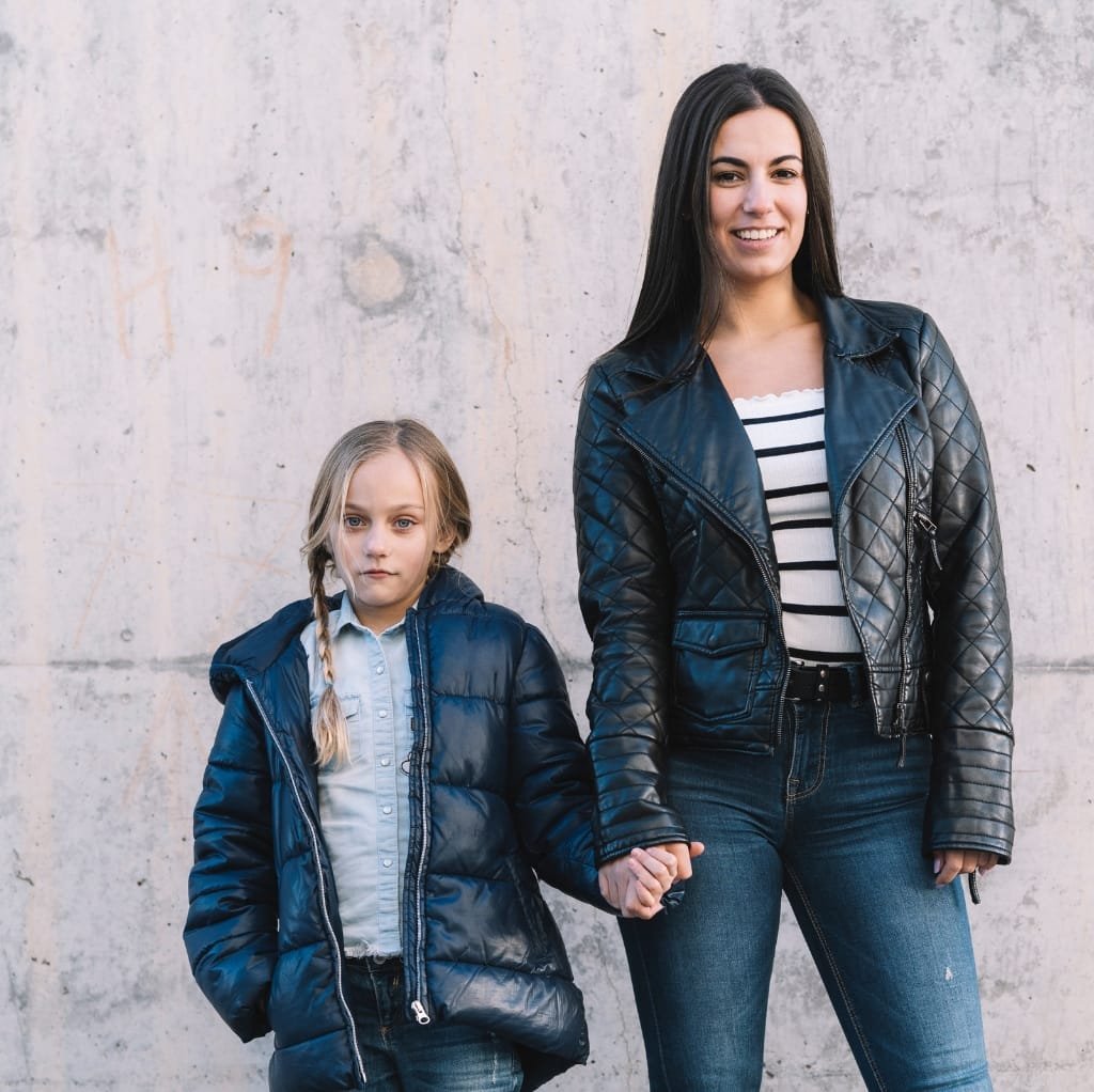 A woman and a young girl standing against a concrete wall, both wearing quilted bomber jackets and holding hands.