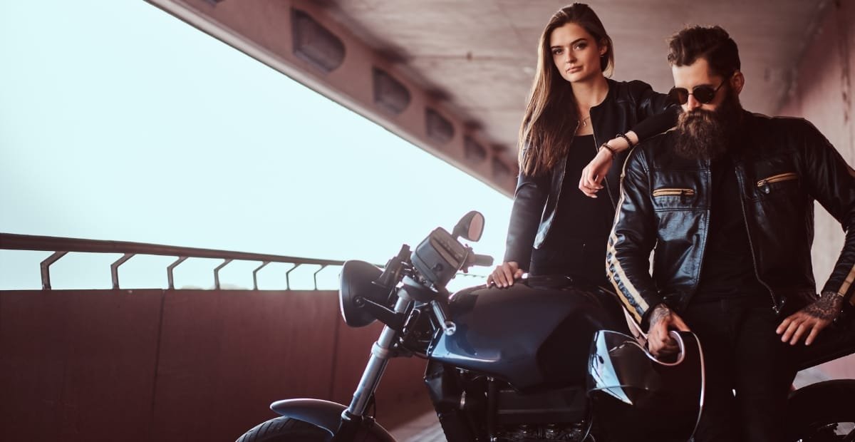 A young woman and a young man with a beard and sunglasses posing next to a motorcycle in a parking garage or underpass.