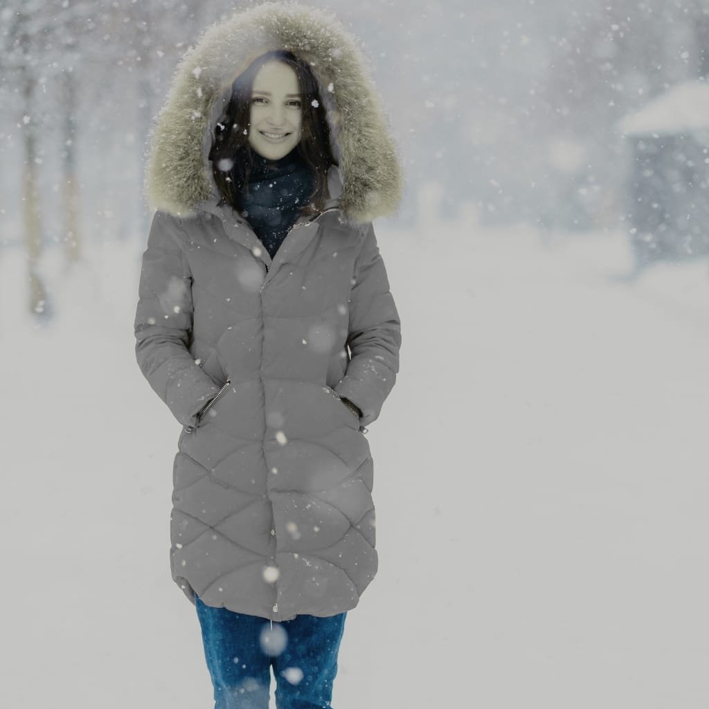 A woman standing outdoors in a snowy winter landscape, wearing a long, grey, insulated winter coat with a fur-lined hood. Snow is falling around her, and she is smiling with her hands in her coat pockets.

