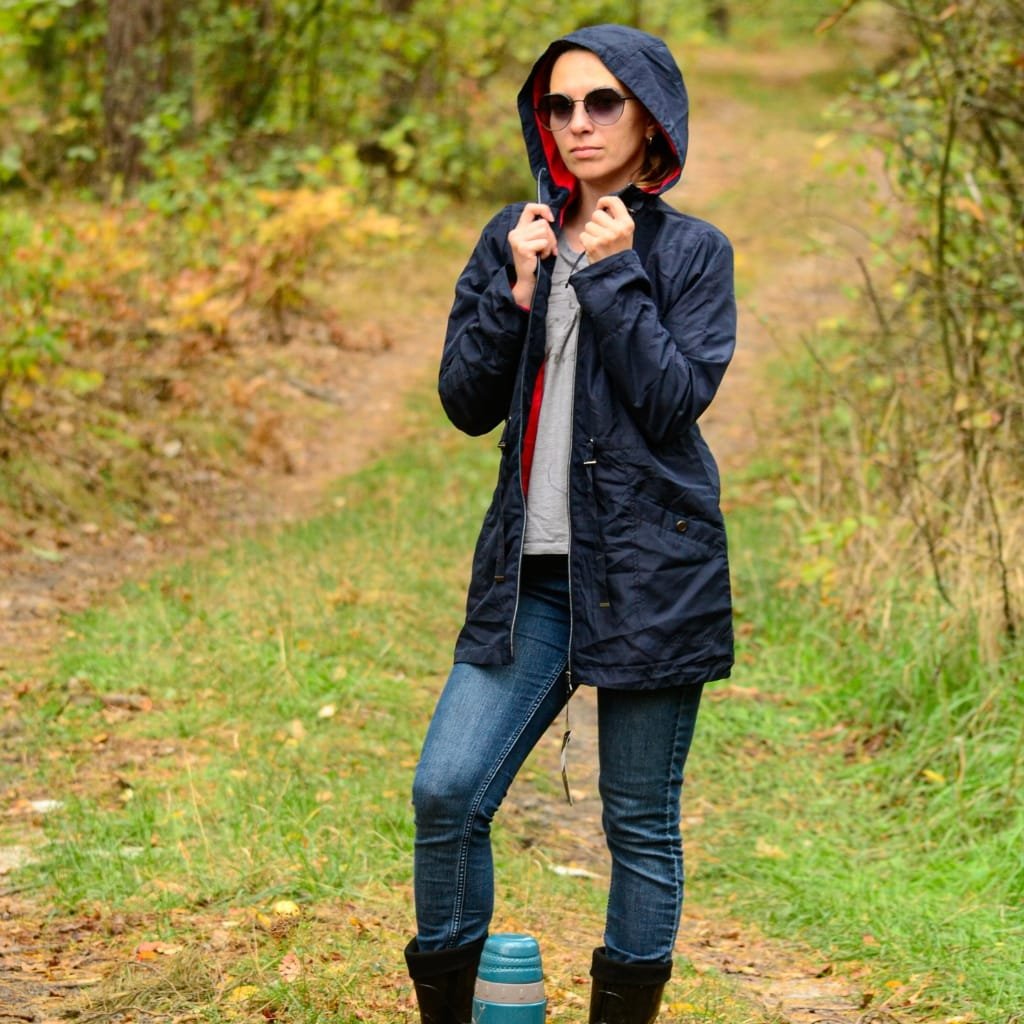 How to Style an Anorak Rain Jacket for Every Occasion 8 A woman standing on a forest trail during autumn, wearing a navy blue anorak rain jacket with the hood up, sunglasses, jeans, and black boots.