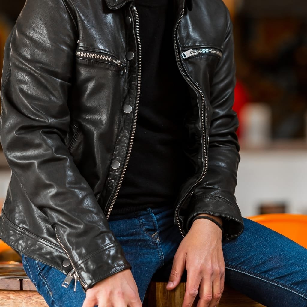 Top Racer Jacket Looks That Never Go Out of Style 8 Close-up image of a person wearing a black leather slim-fit racer jacket, seated with one hand resting on their thigh. The jacket features zipper details on the chest pockets and a front zipper closure, paired with casual blue jeans.