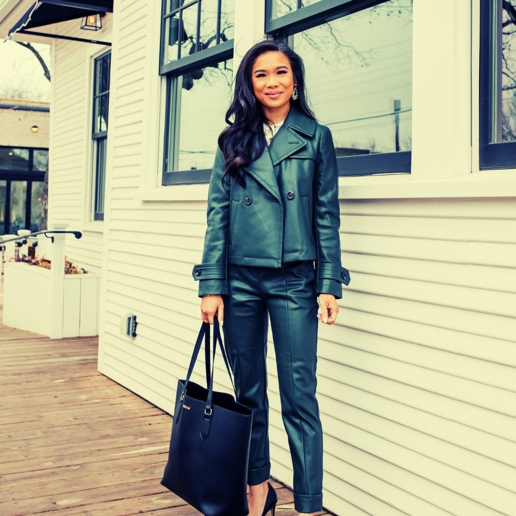 A woman with long dark hair and earrings standing outside, dressed in a dark leather jacket and matching pants, holding a large black handbag. She is smiling and posing in front of a light-colored building with large windows.