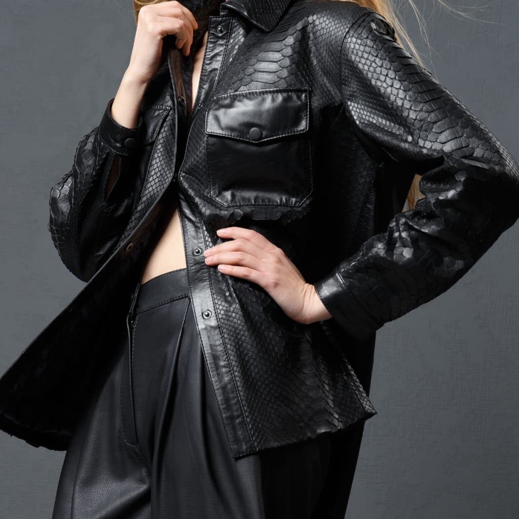 A close-up of a person wearing a black leather jacket with a snap or button closure. The person's hand is resting on their hip, and the jacket features a textured, snakeskin-like pattern.