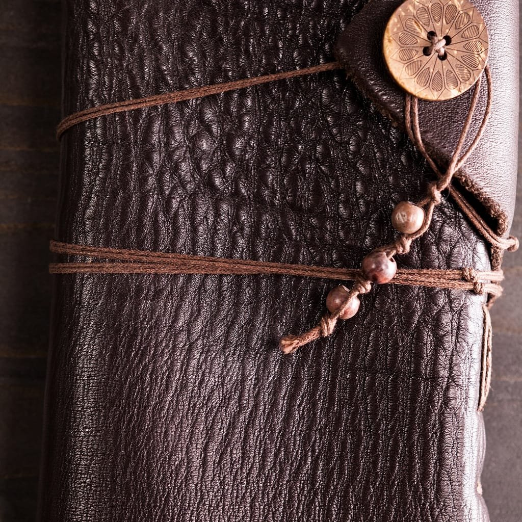 Close-up of a rolled piece of dark brown Split Buffalo Leather wrapped with a brown cord, featuring a decorative button with intricate cut-out patterns and small beads attached by a string.