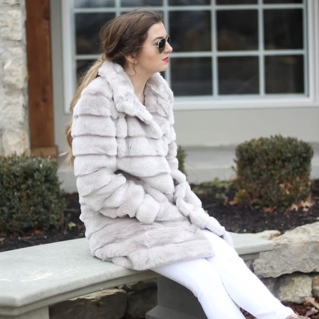 A woman sitting on a stone bench outdoors, wearing a soft, light-colored faux fur coat, white pants, and sunglasses, enjoying a cool spring day.