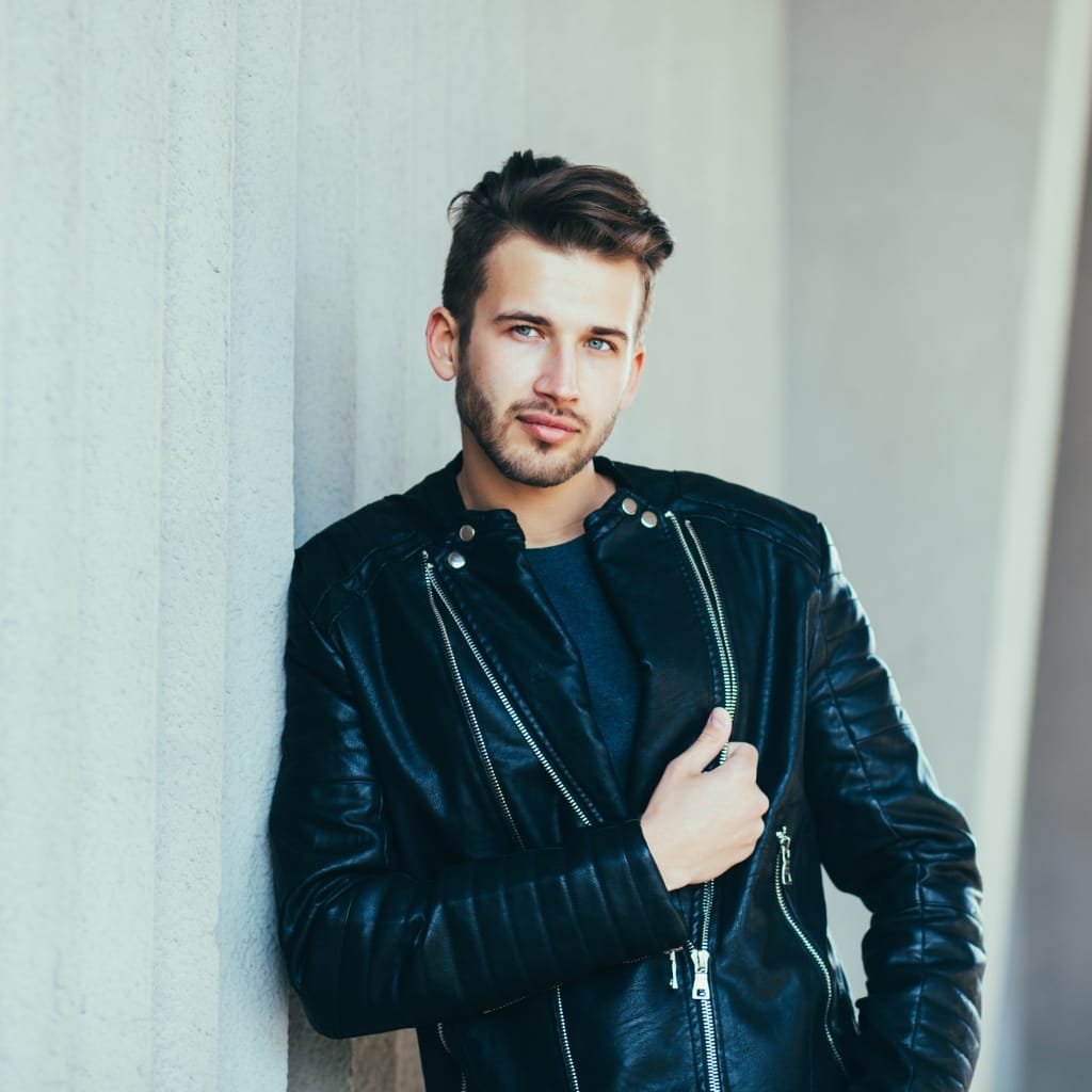 A young man with styled hair and a beard, wearing a black leather jacket over a dark shirt, leaning against a light-colored wall and looking confidently at the camera.

