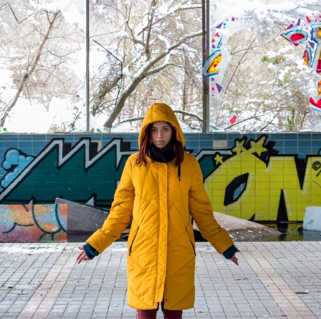 A woman wearing a bright yellow long windbreaker jacket with a hood, standing outdoors in a cold, snowy environment with graffiti-covered walls behind her.