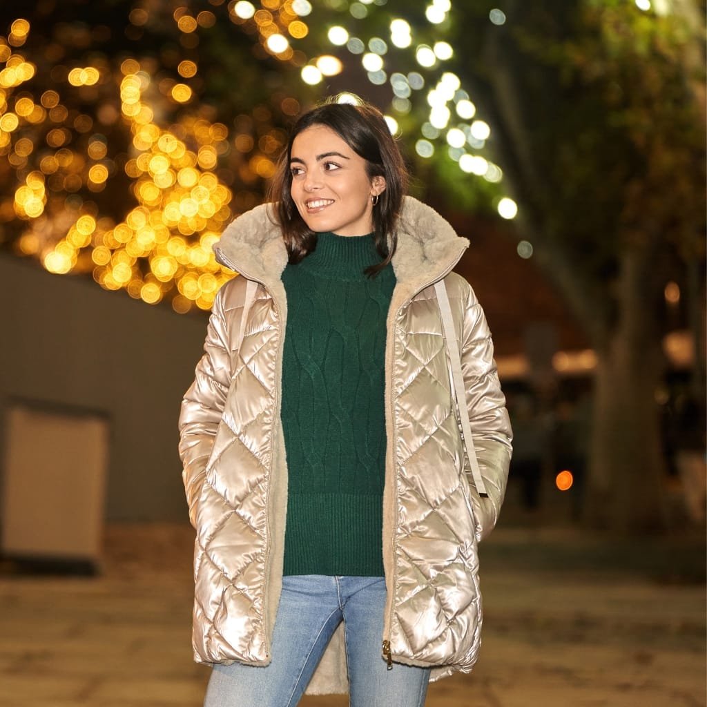 A young woman standing outdoors at night, smiling, wearing a stylish shiny quilted jacket over a green sweater and jeans, with blurred festive lights in the background.


