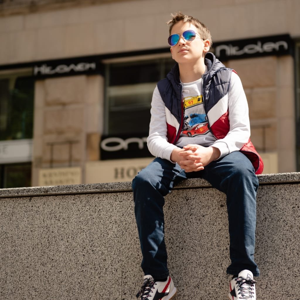 How to Style an Anorak Rain Jacket for Every Occasion 5 A young boy sitting on a stone ledge in an urban setting. He wears reflective sunglasses, a stylish puffer vest with red, white, and navy colors, a graphic t-shirt featuring cars, dark jeans, and Nike sneakers. The background showcases city buildings and signage.