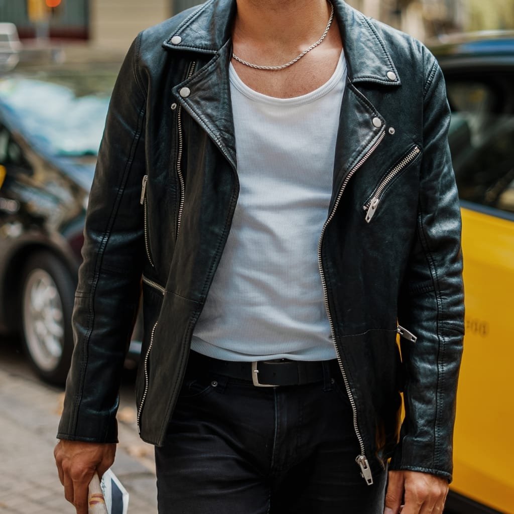 A person wearing a black buffalo leather jacket, a white ribbed T-shirt, black jeans, accessorized with a silver chain necklace and a belt, standing outdoors.