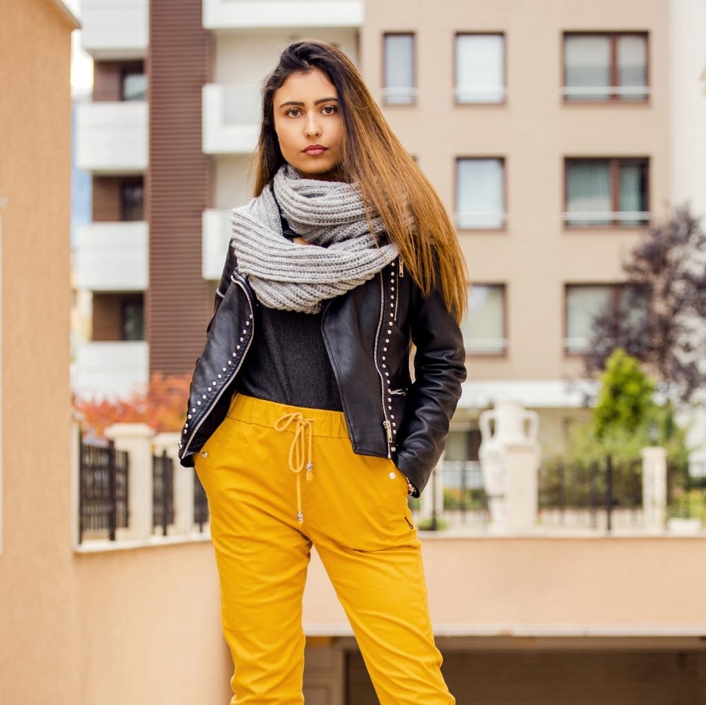 A young woman standing outdoors on a balcony, wearing a black minimalist bomber jacket, yellow pants, and a grey striped scarf, with modern apartment buildings in the background.