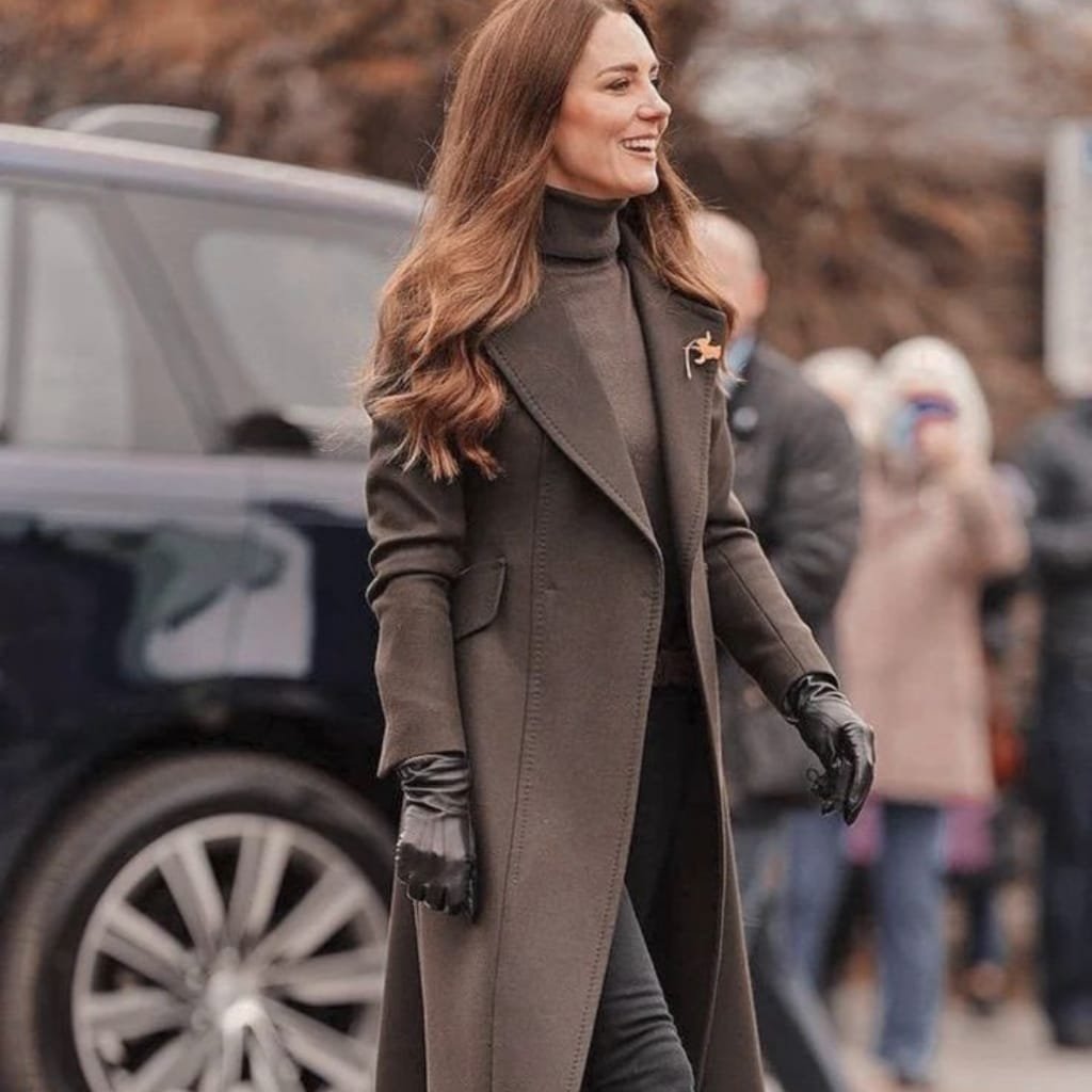 A woman walking outdoors, dressed in a sophisticated dark pea coat layered over a turtleneck, with black gloves. She is smiling and appears to be attending a formal event. The background is blurred, with other people and a vehicle visible.