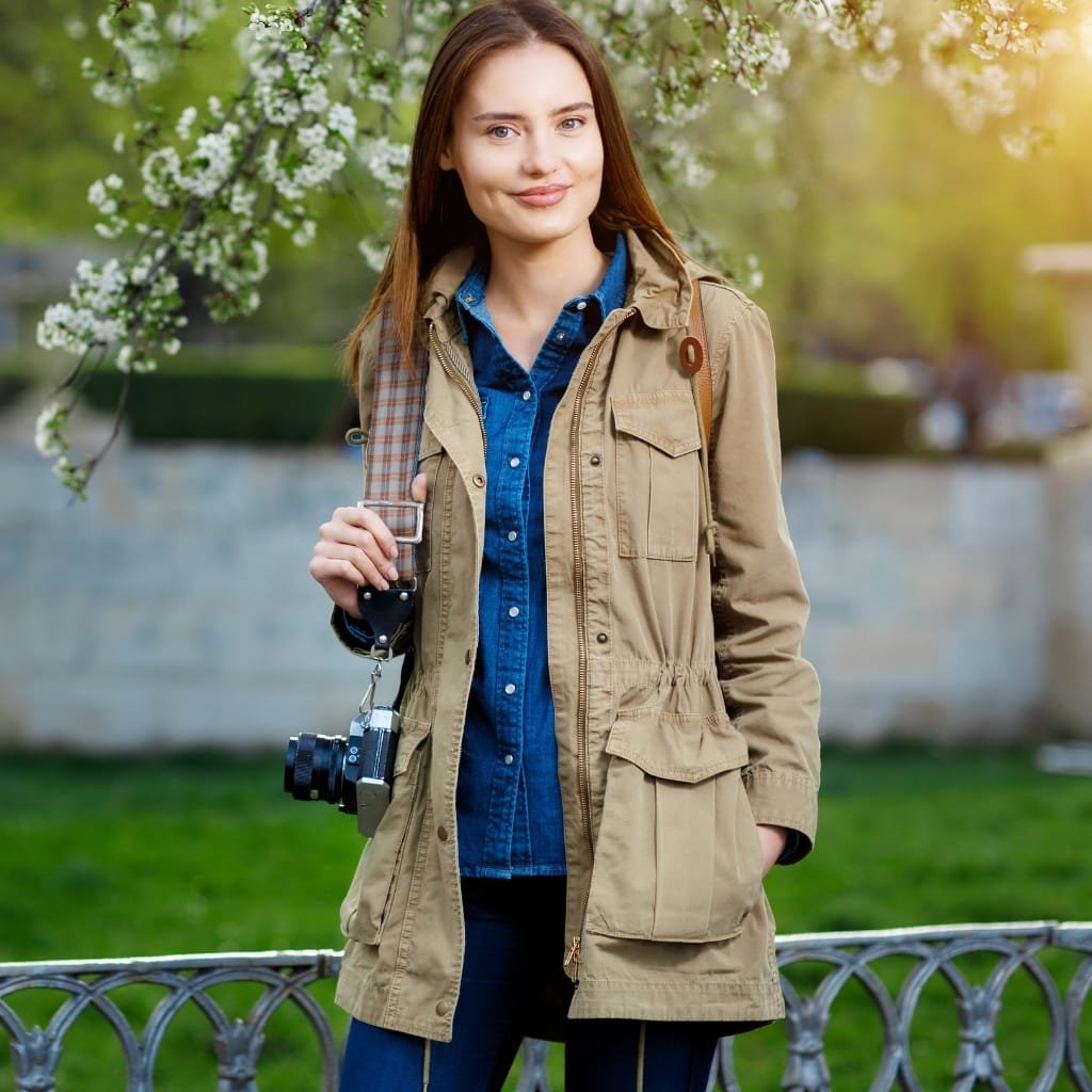 A young woman standing outdoors near a river, wearing a stylish beige lightweight jacket over a denim shirt, with a camera hanging from her neck and a backpack on her shoulder. She is smiling, with blossoming trees and greenery in the background.