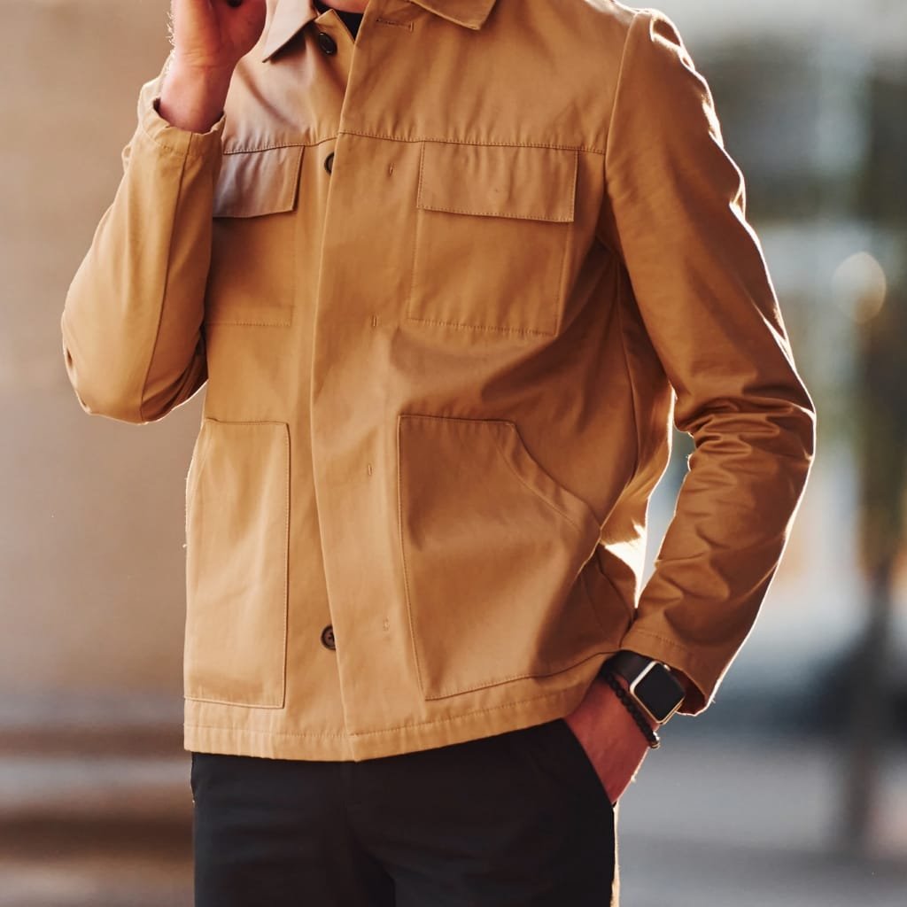 A person wearing a tan suede leather shirt jacket with four front pockets, standing outdoors with one hand in their pocket and the other adjusting their collar.
