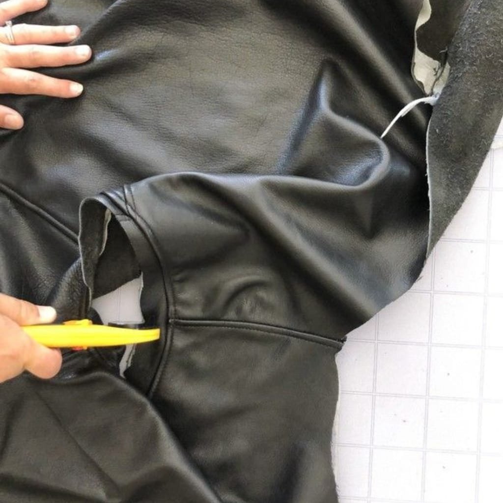 A person is holding a yellow utility knife and appears to be cutting or marking a black cowhide leather shirt jacket, which is laid on a tiled surface. The jacket shows some lines and marks, possibly for tailoring or alteration purposes.