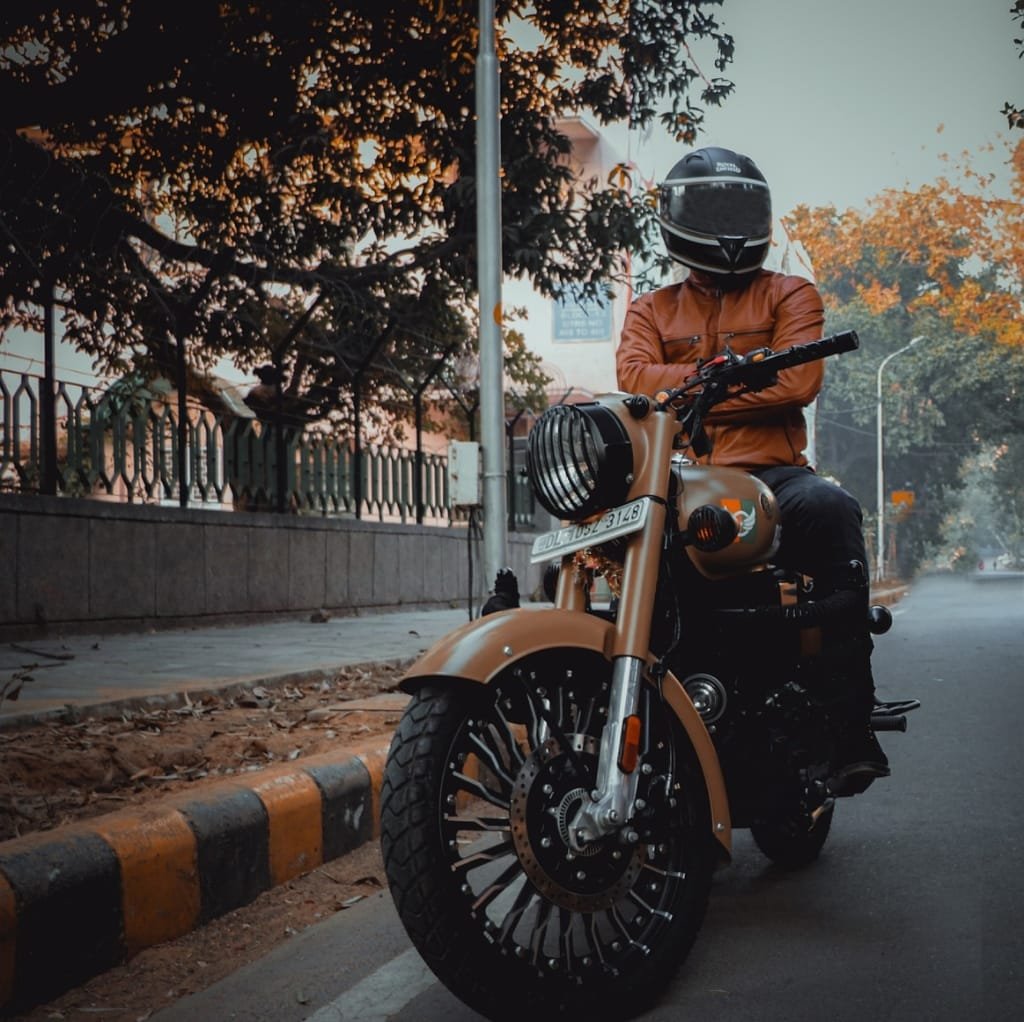 A person wearing a brown leather bomber jacket and a black helmet, sitting confidently on a tan motorcycle on an urban street with autumn trees in the background.