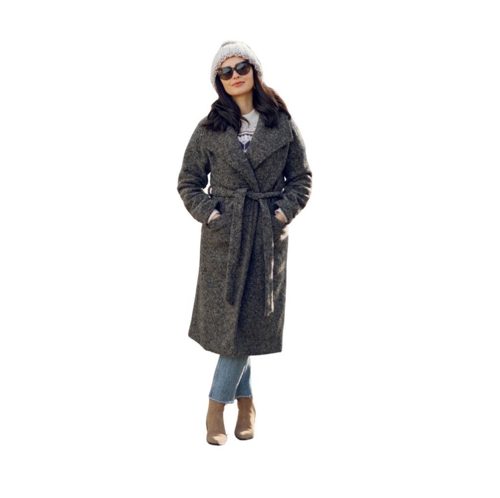 A woman wearing a long, gray wool coat with a belted waist, paired with jeans, beige boots, a knit hat, and sunglasses.
