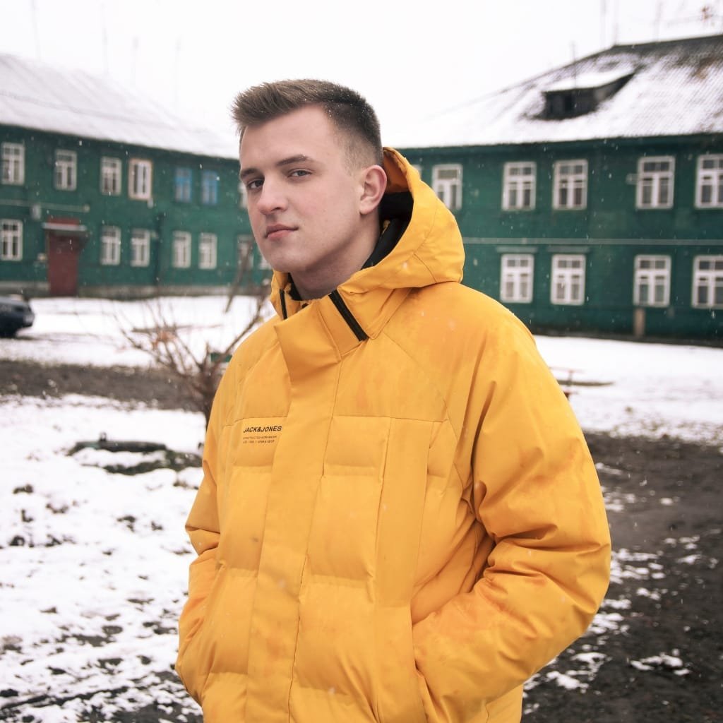 A young man standing outdoors in a snowy area, wearing a bright yellow quilted jacket with his hands in the pockets, with snow-covered ground and buildings in the background.

