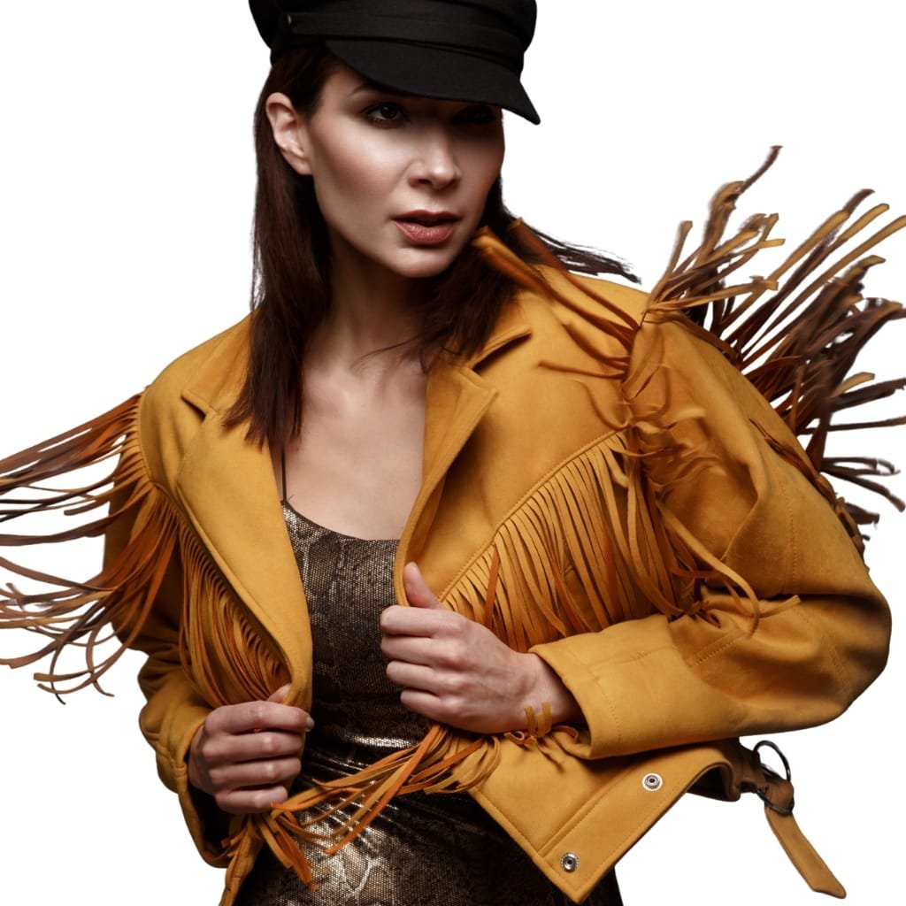 A woman wearing a mustard-colored leather jacket with fringe details on the shoulders and sleeves, paired with a dark hat and a patterned top underneath.


