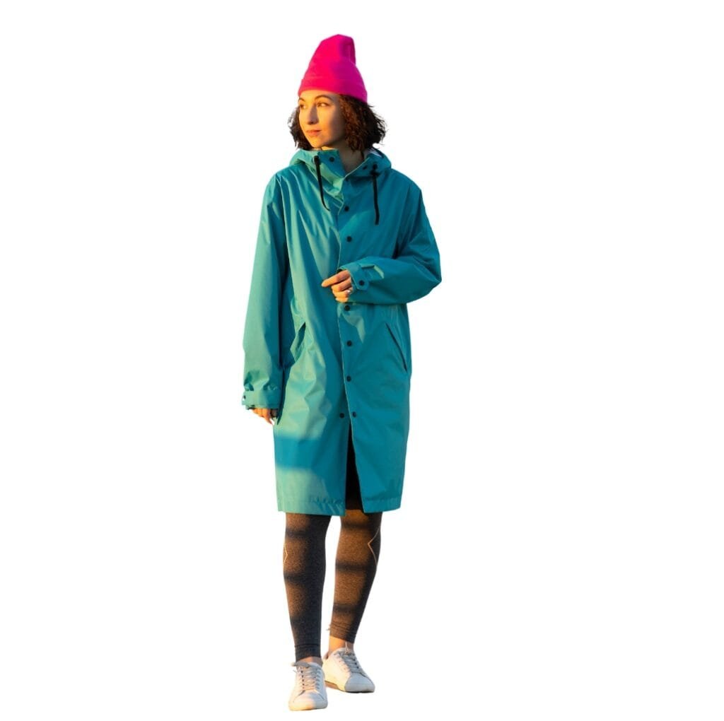 A woman wearing a vibrant teal long windbreaker jacket, a bright pink beanie, patterned tights, and white sneakers, standing outdoors with a confident pose.