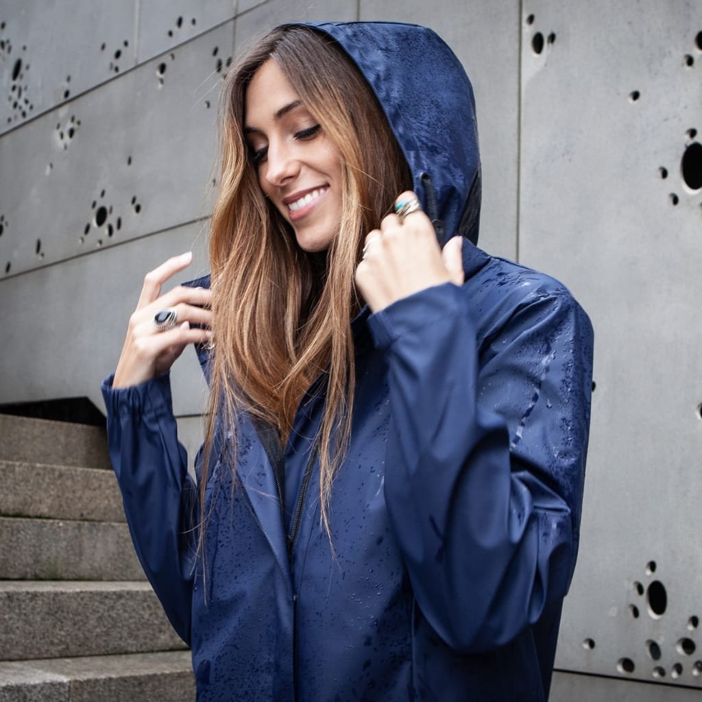 A woman smiling and adjusting the hood of her blue waterproof rain jacket, standing outdoors against a modern concrete wall with circular holes.

