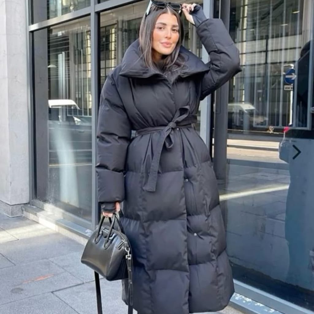 Fashionable woman layering a puffer coat over a hoodie with boots and jeans on a snowy city street.
