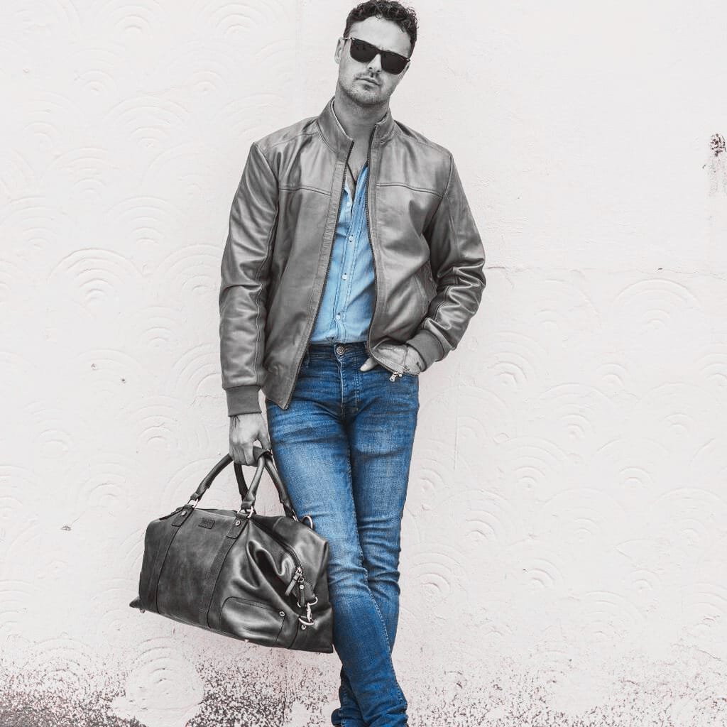 A stylish man with dark hair and sunglasses standing against a textured white wall. He wears a shiny leather jacket over a light blue denim shirt, with matching blue jeans, and carries a large black leather duffel bag.