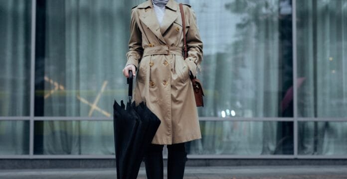 Woman wearing a classic beige trench coat holding a black umbrella in front of glass building.