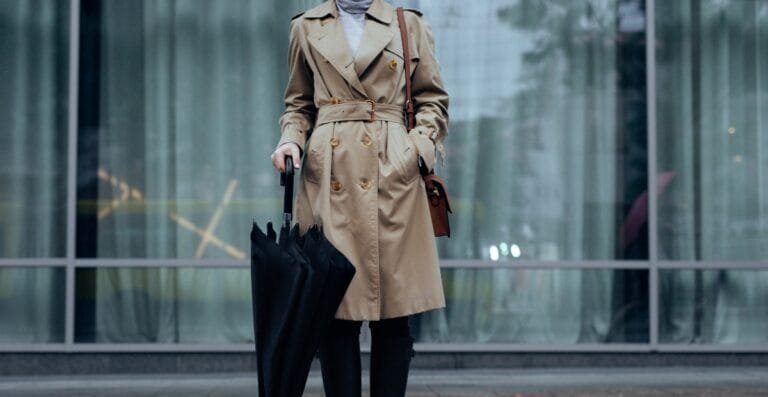 Woman wearing a classic beige trench coat holding a black umbrella in front of glass building.