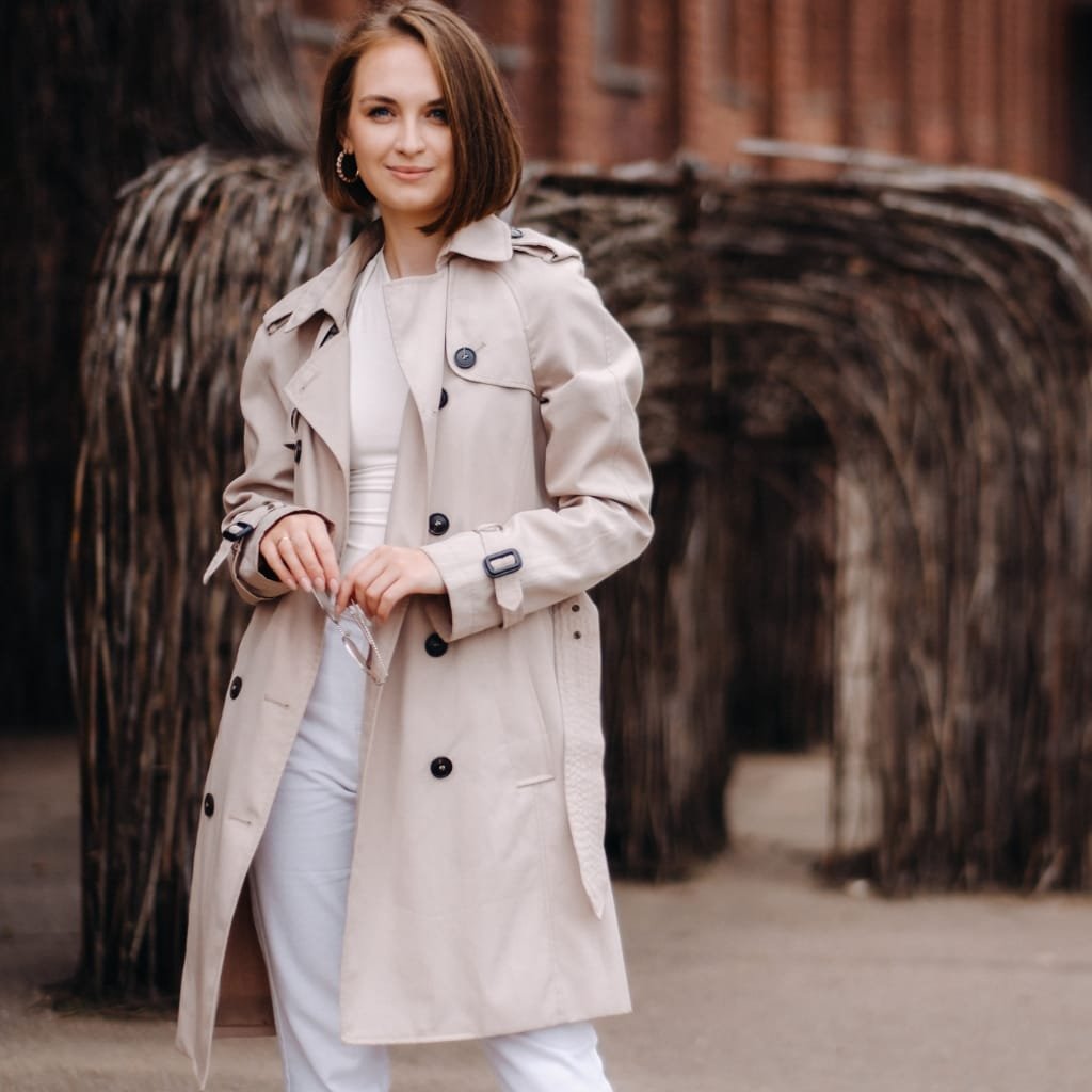 A woman standing outdoors wearing a stylish beige trench coat over a white outfit, with a blurred background of wooden structures and natural elements.
