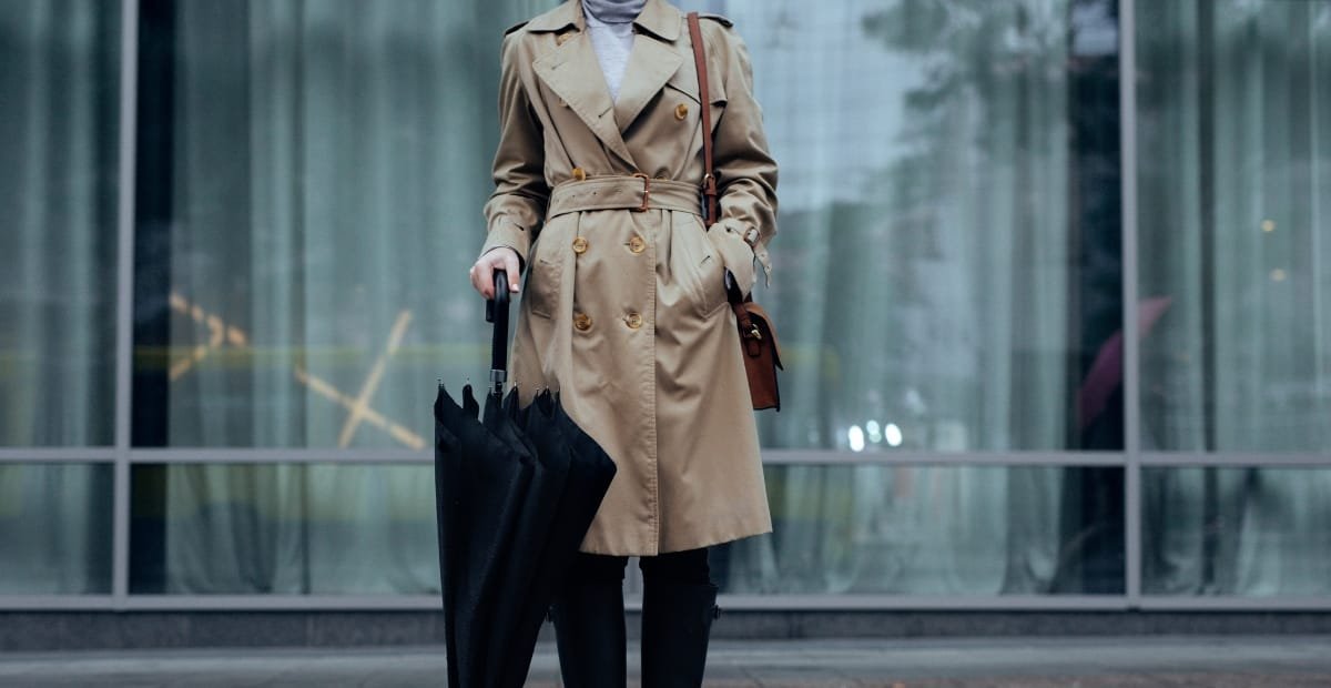 How to Style Trench Coats for Every Season Woman wearing a classic beige trench coat holding a black umbrella in front of glass building.