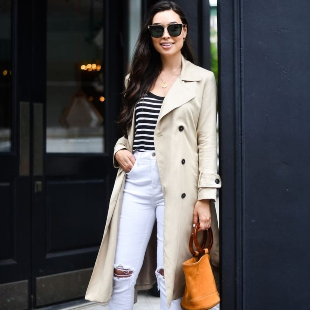 How to Style Trench Coats for Every Season 5 Stylish woman in a beige trench coat with striped top, white ripped jeans, and tan handbag, standing outside.