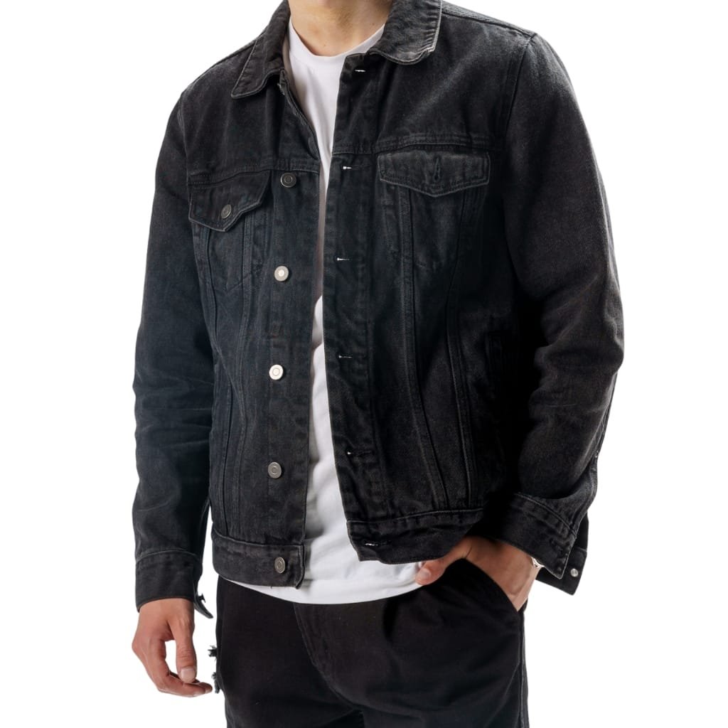 Man wearing a black denim trucker jacket, white T-shirt underneath, and black pants, with one hand in his pocket.