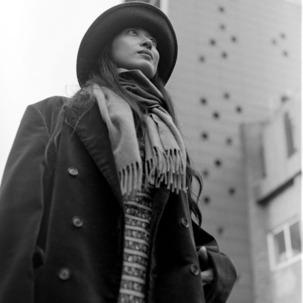 A black-and-white photo of a young woman looking upward, wearing a wide-brim hat, a scarf around her neck, and a pea coat, standing against an urban building background.