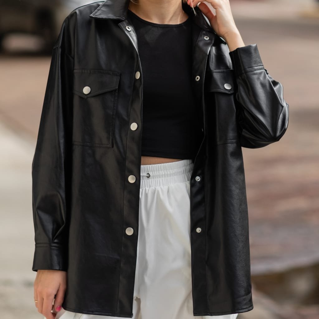 A person wearing a black leather utility shirt jacket over a black cropped T-shirt, paired with white drawstring pants. The jacket features large front pockets with button closures and silver snap buttons along the front.