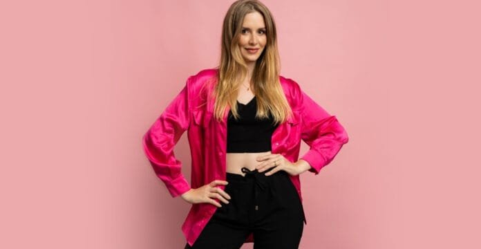 A woman poses against a pink background, wearing a shiny, bright pink velvet bomber jacket over a black crop top and black pants.