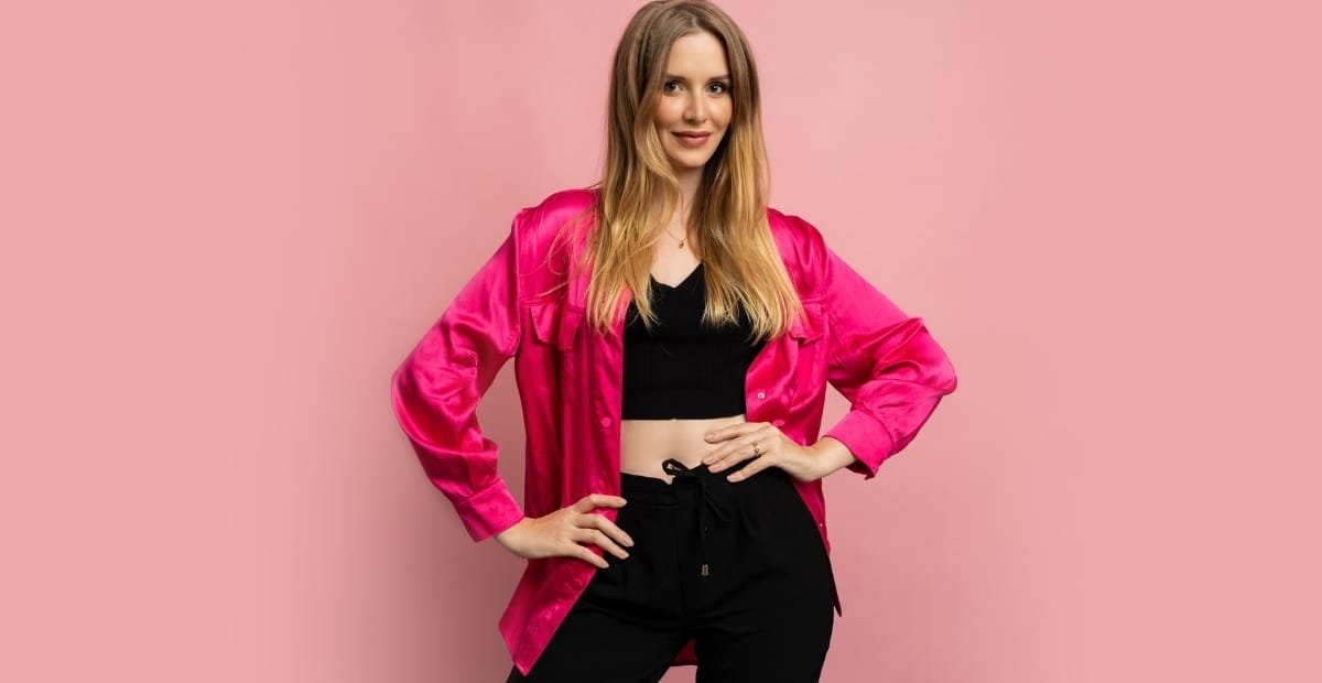 How to Style a Velvet Bomber Jacket for Casual and Luxe Looks A woman poses against a pink background, wearing a shiny, bright pink velvet bomber jacket over a black crop top and black pants.