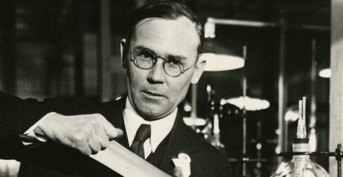 Black and white photograph of a man wearing glasses and a suit, working with a scientific glass apparatus in a laboratory.